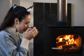 Obraz premium Woman cupping small dark cup at home, leaning toward wood stove in knit sweater, copy space