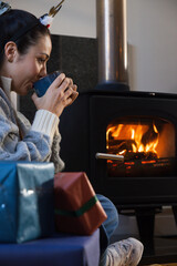 Obraz premium Female sitting by wood-burning stove in living room sweater sipping blue mug near gifts, copy space
