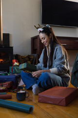 Obraz premium Adult female wearing reindeer headband in living room opening wrapped gifts by stove with mug