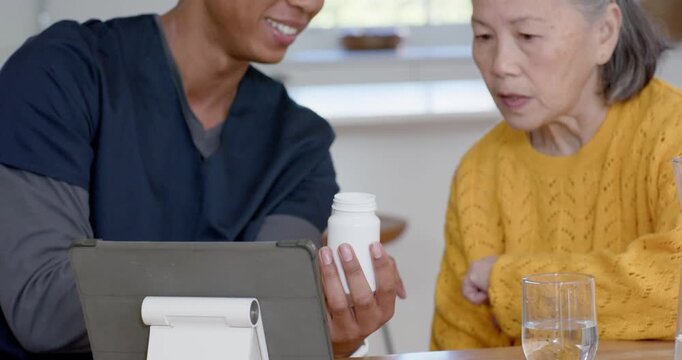 Diverse caregiver helping senior Asian woman at table, reading tablet showing bottle for dose