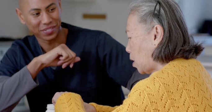 Diverse aide in scrubs guiding Senior Asian woman handling pill bottle, explaining meds at kitchen