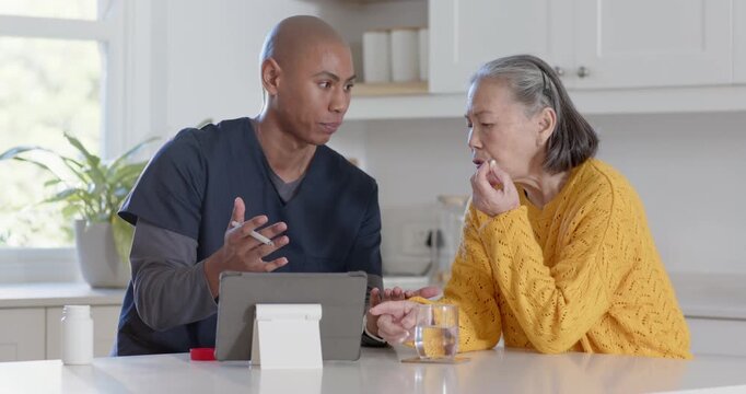 Nurse in scrubs pointing stylus explaining dose on tablet in kitchen as senior woman holding pill