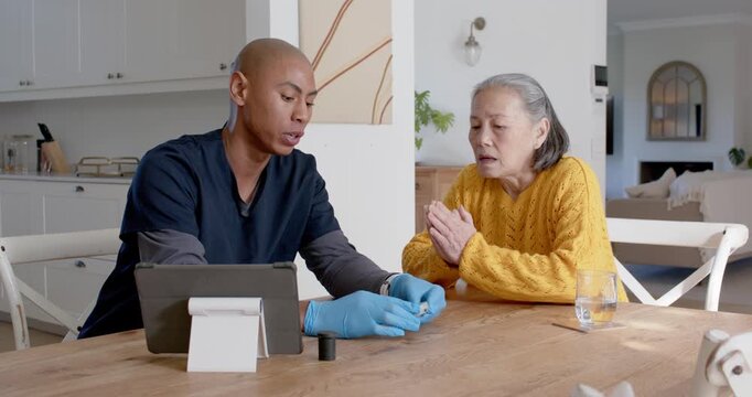 Caregiver in scrubs picking strip, teaching meter and tablet use to senior Chinese at table