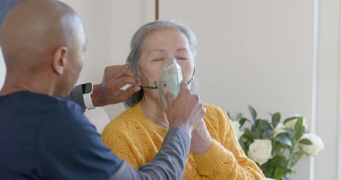 Caregiver assisting Senior Asian woman after closing eyes, fixing O2 mask on couch in living room
