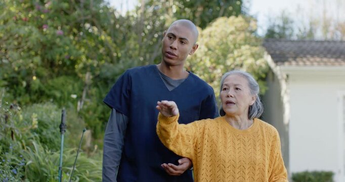 Senior Asian woman holding cane on path pointing to house, African-American navy-clad nurse guiding