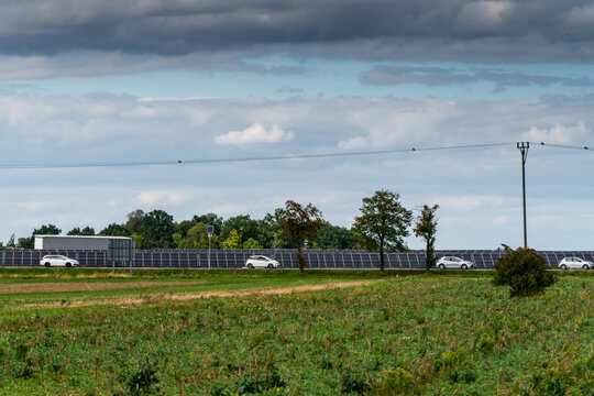View of a solar farm with rows of photovoltaic panels situated along a road with passing cars in a rural landscape under a cloudy sky in Poland.