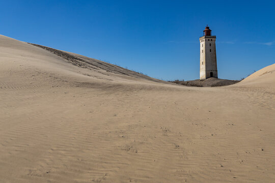 View of the historic Rubjerg Knude lighthouse standing amidst vast sand dunes under a clear blue sky in Lokken, Jutland, Denmark.