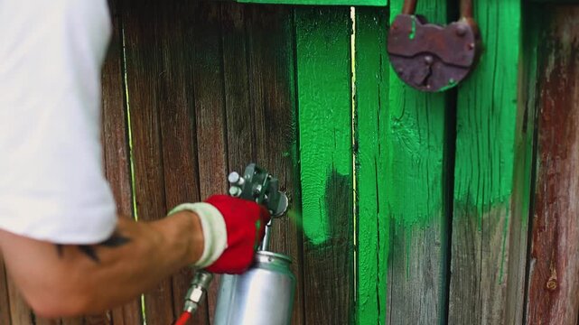 Male worker using air compressor to spray paint a green wooden gate in a garden, wearing gloves and casual clothing, with grass and wall visible in the background	
