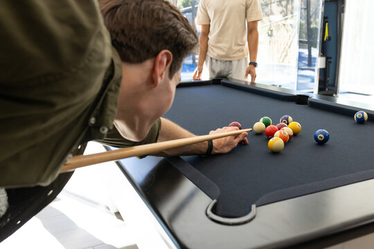 Two males aiming over black pool table in lounge holding cue with clustered balls, copy space