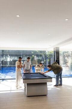 Diverse male friends playing pool, leaning over blue felt table in recreation room, holding cues