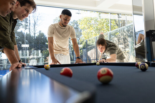 Diverse male friends casually dressed playing pool with cues and balls in rec room, copy space