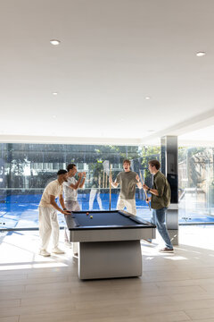 Diverse male friends playing at pool table in modern game room with cues and glass wall