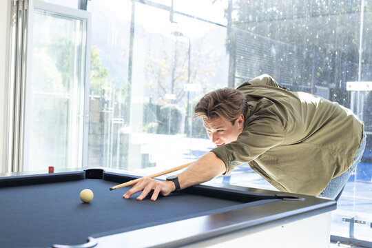 Adult man lining up shot on black-felt pool table in glass-walled lounge, aiming at white ball