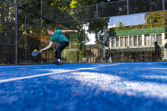 Adult male reaching low, hitting yellow padel ball with perforated racket on padel court