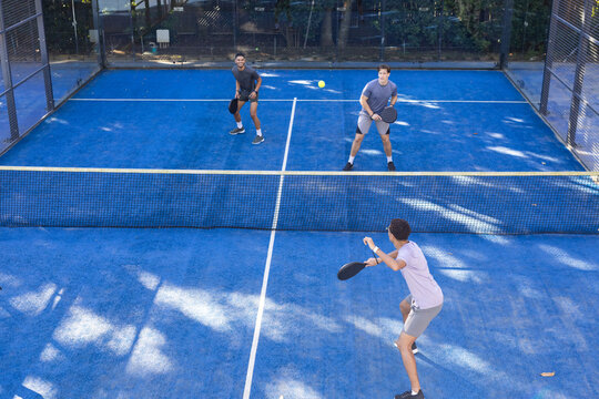 Diverse male friends playing paddle tennis on blue outdoor court, swinging paddles, yellow ball