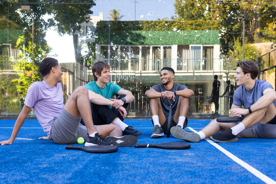 Four men sitting on blue padel court wearing athletic t-shirts and shorts, holding paddles and ball