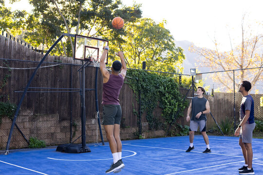 Diverse male friends jumping and shooting basketball on backyard blue court with portable hoop