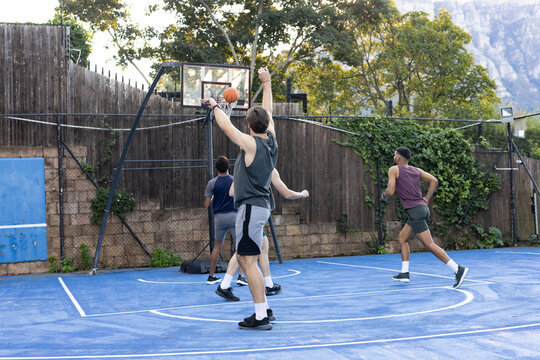 Diverse male teammates playing basketball on blue court, shooter rising, bag at hoop, in shorts