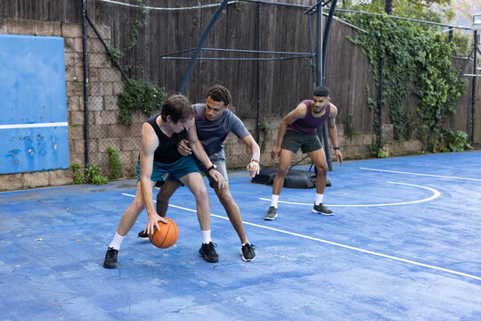 Diverse male friends wearing tanks playing pickup basketball on blue court, dribbling orange ball