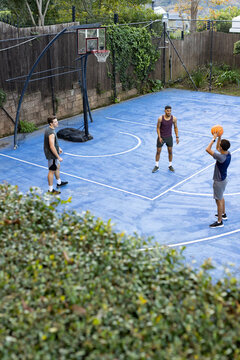 Diverse male friends playing on blue yard court holding orange basketball near portable hoop, fence