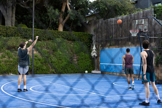 Diverse male friends shooting basketball on bright blue backyard court with hoop
