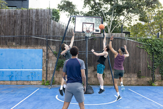 Diverse male friends in shorts playing basketball on backyard court, shooting at hoop and guarding