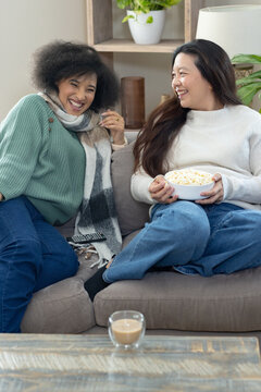 Diverse female friends laughing and chatting on gray sofa in living room with popcorn bowl, remote