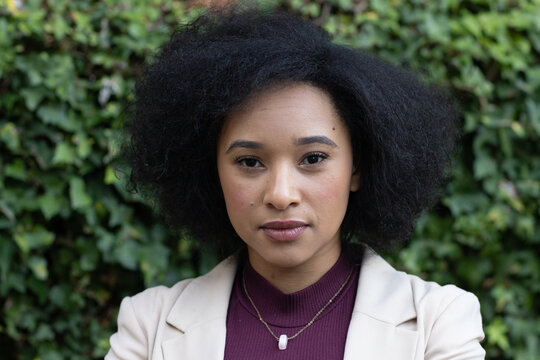 African American woman posing by ivy wall beige blazer, burgundy top, gold chain with white pendant