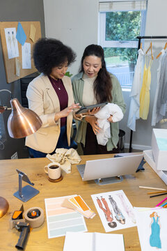 Diverse female colleagues pointing at tablet and examining bundled fabric at design studio desk