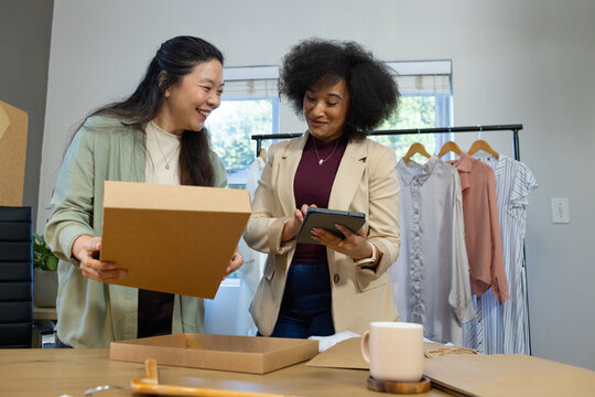 Diverse female coworkers packing orders and checking tablet at small studio worktable with garments