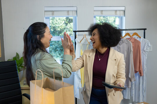 Female colleagues high-fiving and laughing in shop by garment rack, holding tablet and shopping bag