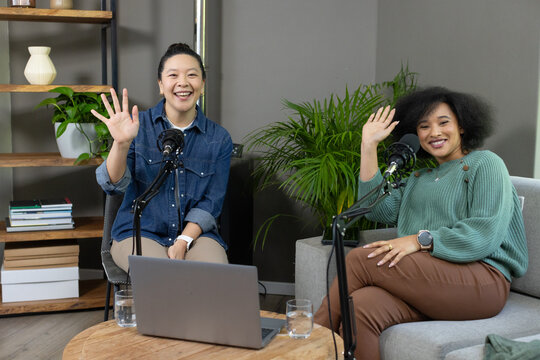 Diverse female podcast hosts waving at camera while sitting in home studio with mics and laptop