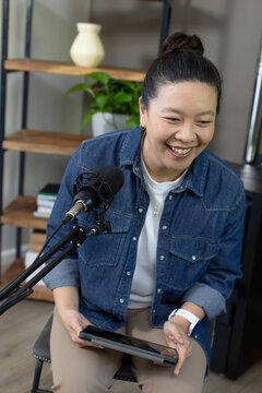 Asian woman smiling, leaning toward boom microphone while holding tablet in home office