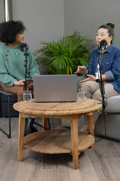 African American and Asian women co-hosts talking and gesturing at coffee table with mics laptop
