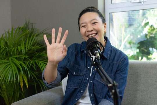 Asian woman sitting on grey sofa wearing denim shirt, waving hand at microphone, smiling