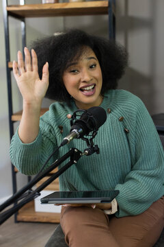 African woman sitting in studio in green sweater brown pants, speaking into mic and holding tablet