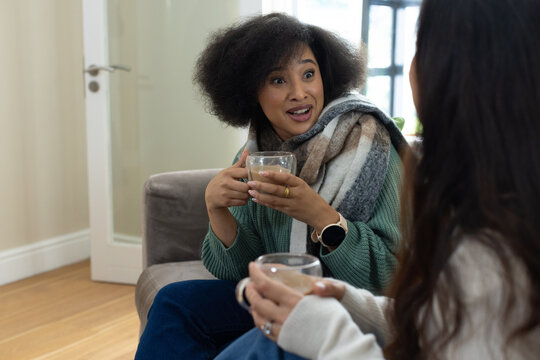 Diverse female friends sitting on gray sofa at home, wearing patterned scarf and holding glass cups