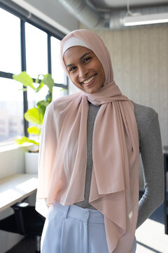 African American woman standing by large window in office, wearing pale pink hijab beside plant