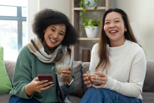 Diverse female friends sitting on gray sofa at home holding phone and mugs, wearing green sweater