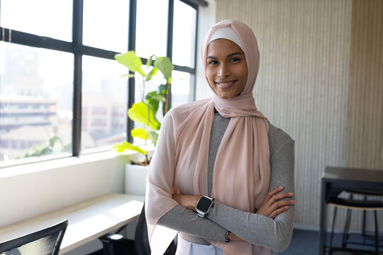 Female wearing pale pink hijab and grey top, standing by large window showing smartwatch and plant