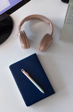 Blush-pink over-ear headphones resting on white desk, navy notebook and silver laptop visible