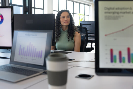 African American woman wearing sleeveless top at desk reviewing laptop, monitor charts with cup