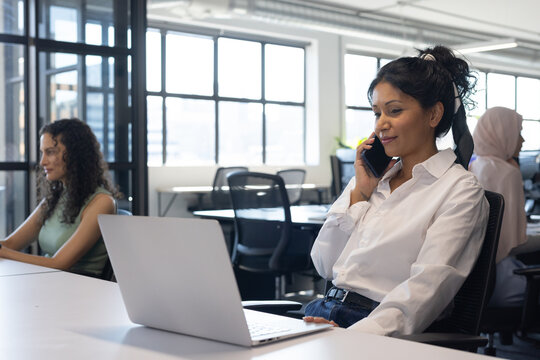 Female coworkers in business attire juggling at desks in open office with laptop, smartphone