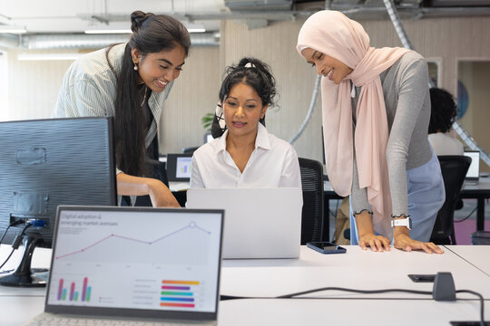 Diverse female coworkers collaborating at office laptop, wearing business attire, pointing at chart