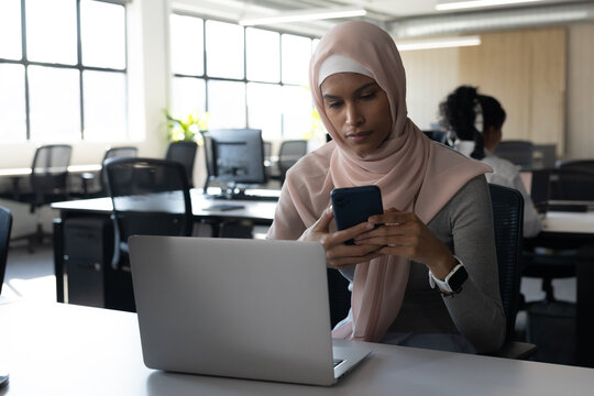 Female coworkers in modern office, African American woman in pink scarf using smartphone and laptop