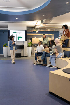 Diverse coworkers in biz-casual presenting and taking notes in office lounge with screen, mic, pad