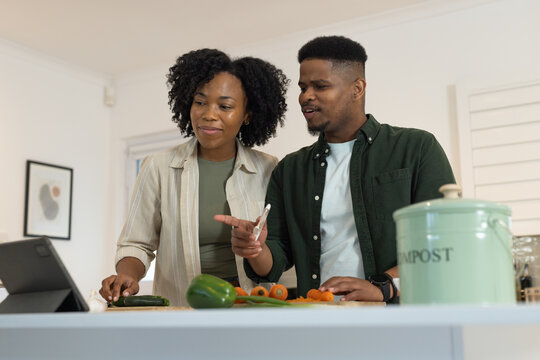 African American couple referencing tablet, slicing cucumber on cutting board in kitchen, in green