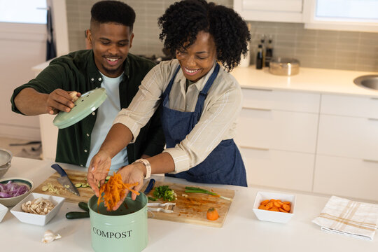 African American couple wearing apron prepping veggies, composting in COMPOST bin on kitchen island