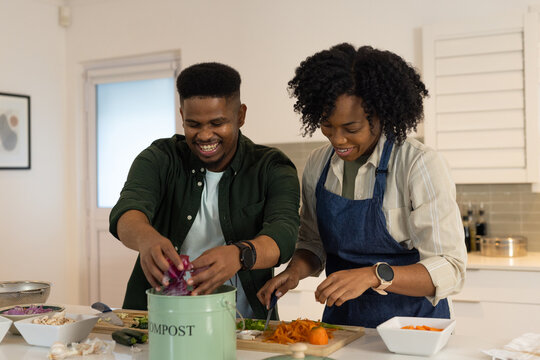 Couple cooking at island, man adding scraps to compost bin, woman in denim apron chopping