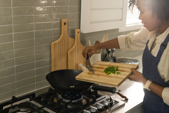 African woman wearing apron adding onions, garlic into wok in kitchen over blue flame, copy space